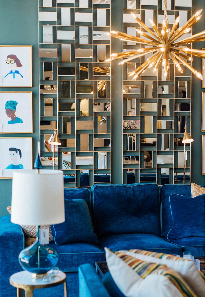 A seating area with a blue couch, mirrored wall decor behind it with framed images next to it, and a chandelier 