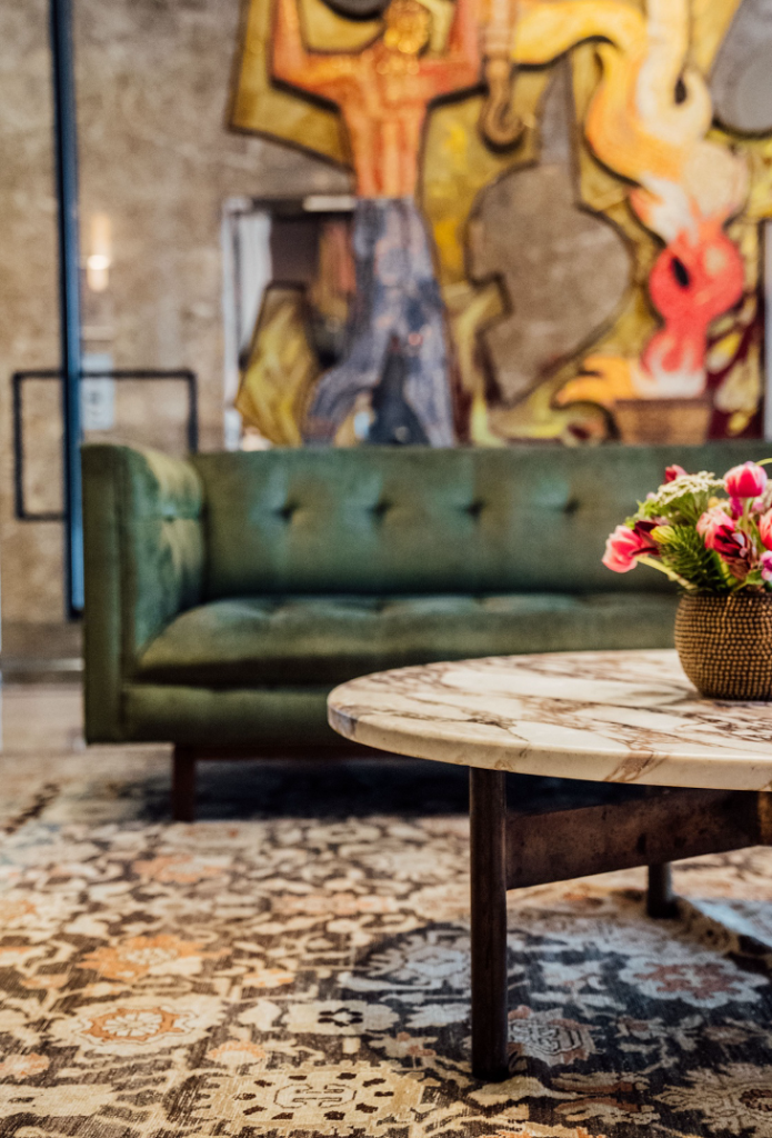 A seating area with a green couch, and eclectic rug, a circular table with  flowers, and an a piece of abstract art in the background