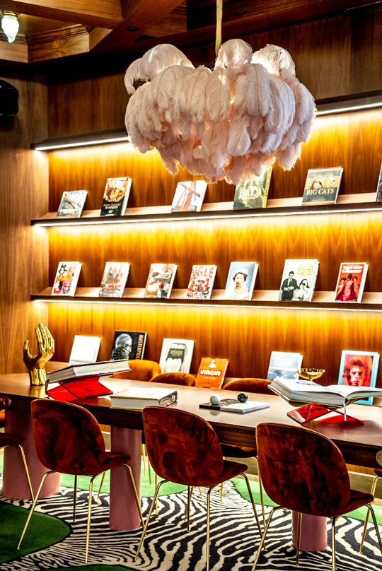 A moody room with a long table with red, velvet chairs. Behind the table is a wall of shelves that are full of books. There is a zebra skin rug below and a feather chandelier above
